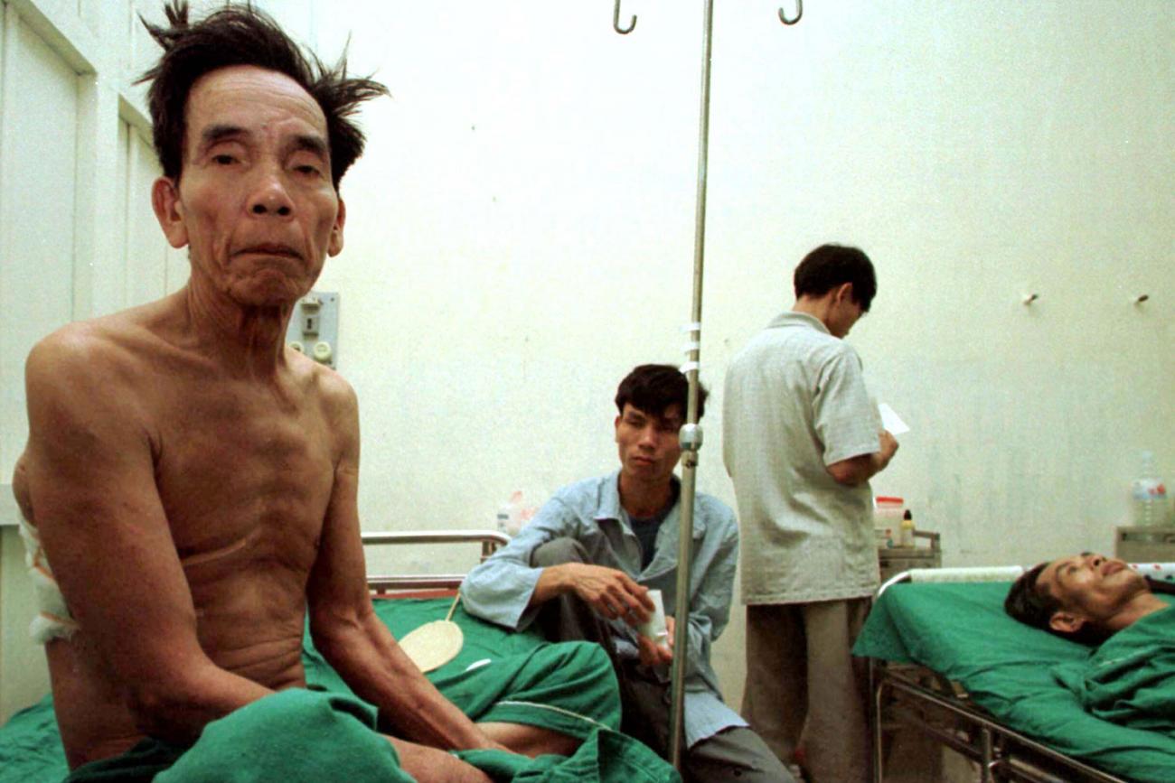 In the picture the man sits with no shirt facing the camera as another patient can be seen lying in the bed in the background. One medical staff is nearby, and a young man sits by the bedside.