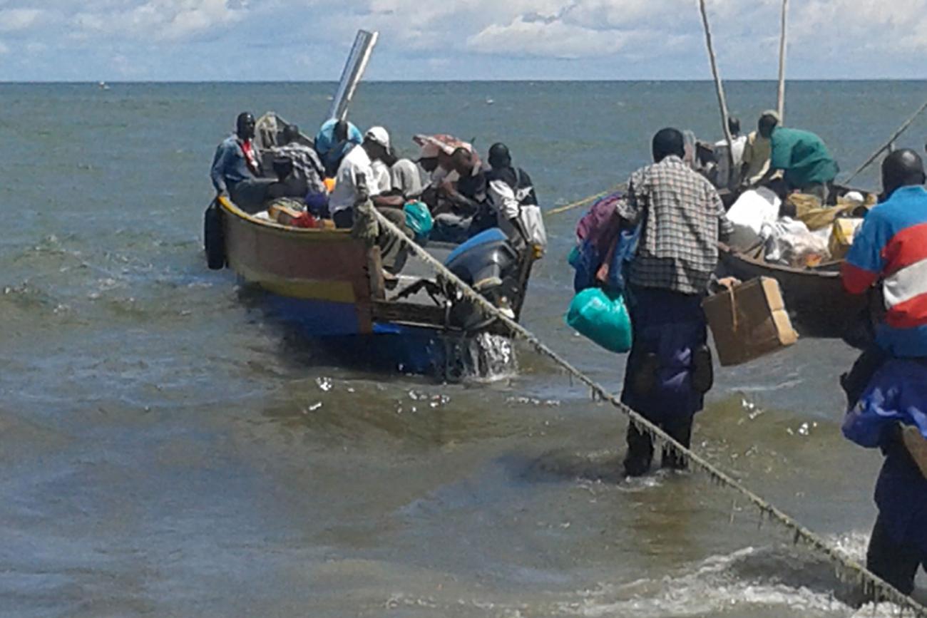 A small boat on the shore of Lake Victoria in Uganda teeming with passengers. The picture shows two boat, one filled with passengers and one with what appears to be lots of small package cargo. Both are tied at the stern to something off camera, and a man with his arms full of bundles wades through knee-high water making his way onto one of the boats.
