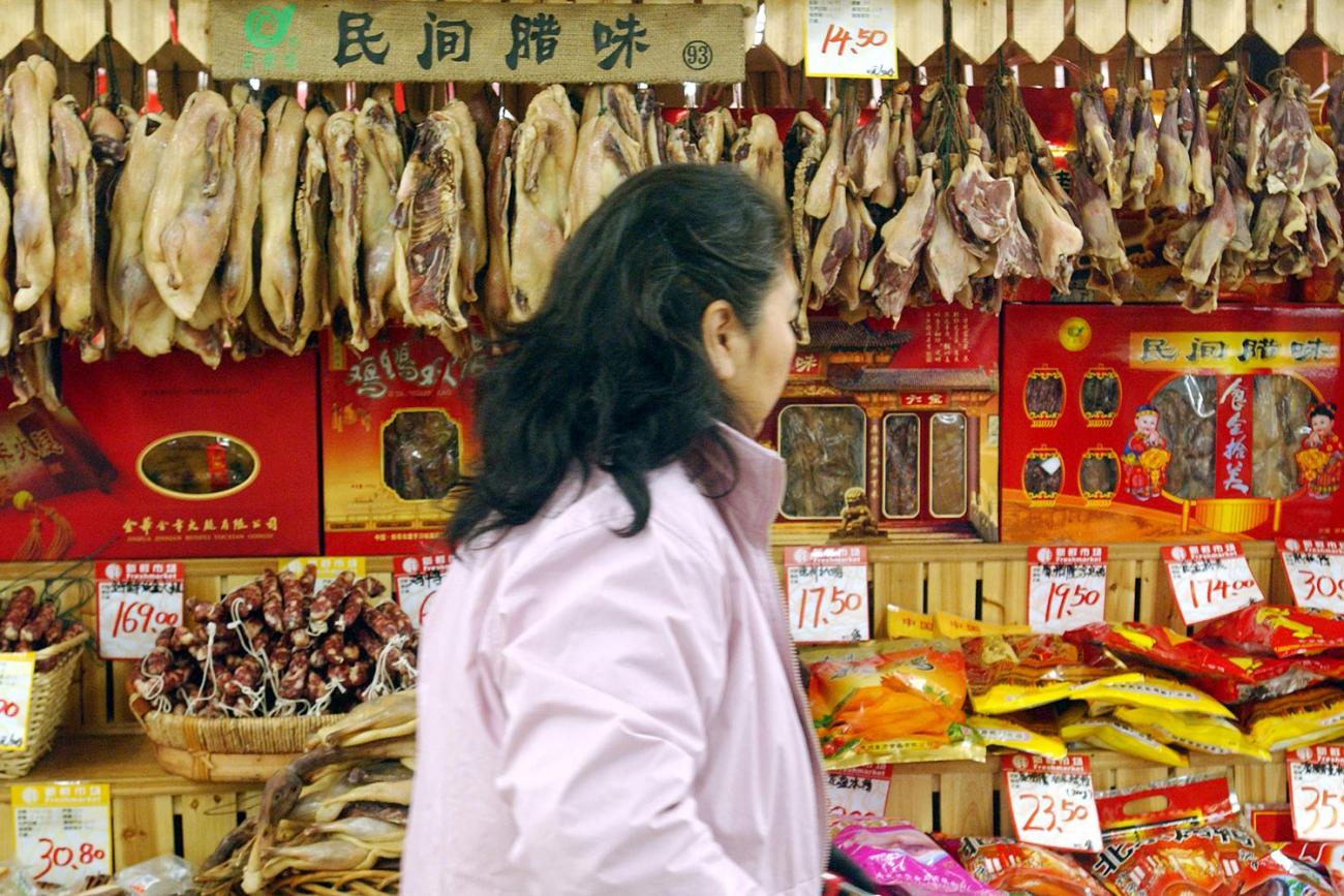Walking by the camera is an older woman with her hair pulled back wearing a dull pink jacket. She walks perpendicular to the direction of the camera and is looking away, at the various foods on display. Behind her is a dizzying array of red- and yellow-hued dried meats and fishes, sausages and packages.