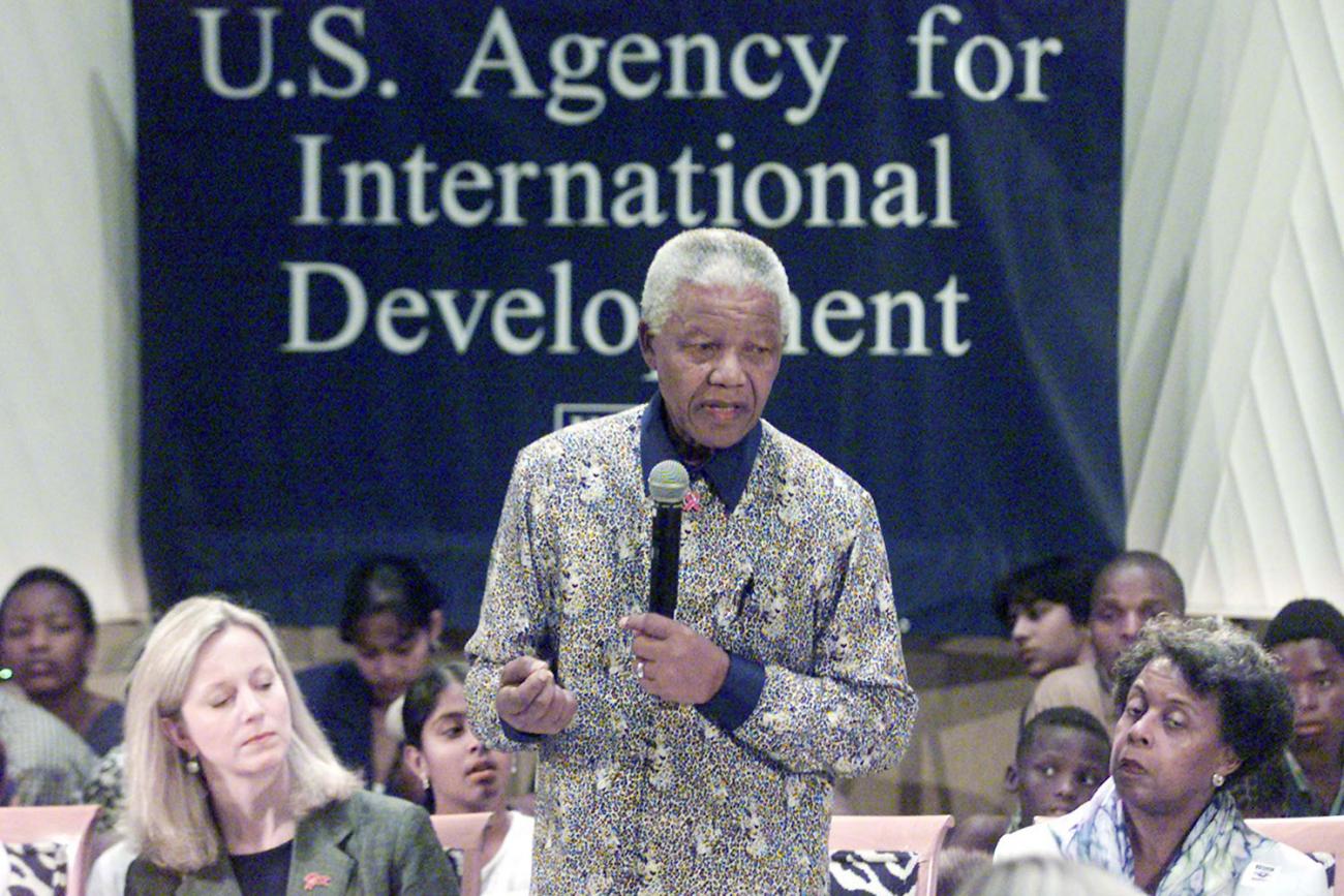 Image shows Nelson Mandela speaking into a microphone before a sign that reads, “U.S. Agency for International Development” at the International AIDS Conference in Durban, South Africa on July 14, 2000. He is standing and is surrounded by people who are sitting and appear to be listening intently.