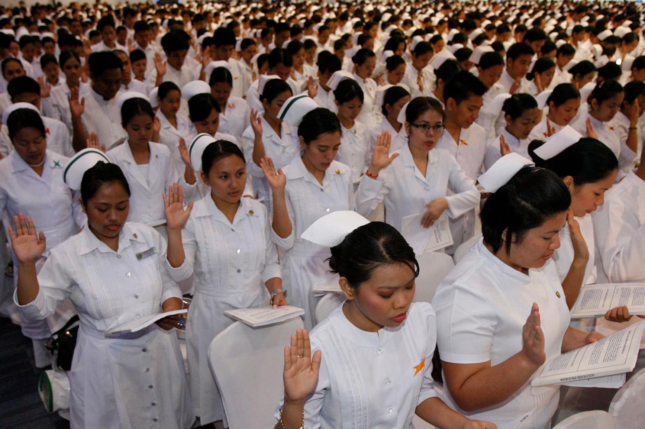 Picture shows thousands and thousands of nurses in uniform lined up with right hands raised reading from a thick packet. The women have traditional nurse caps, but the men's heads are bare.