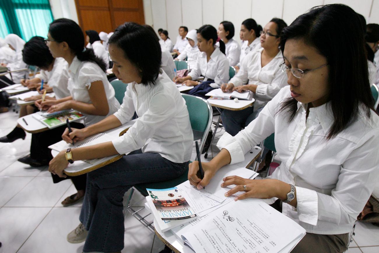 Picture shows a classroom with several rows of nurses at desks, all with white shirts and black pants. They are taking notes and one of them in the foreground has a book on her desk titled, "Japanese Phrase Book."