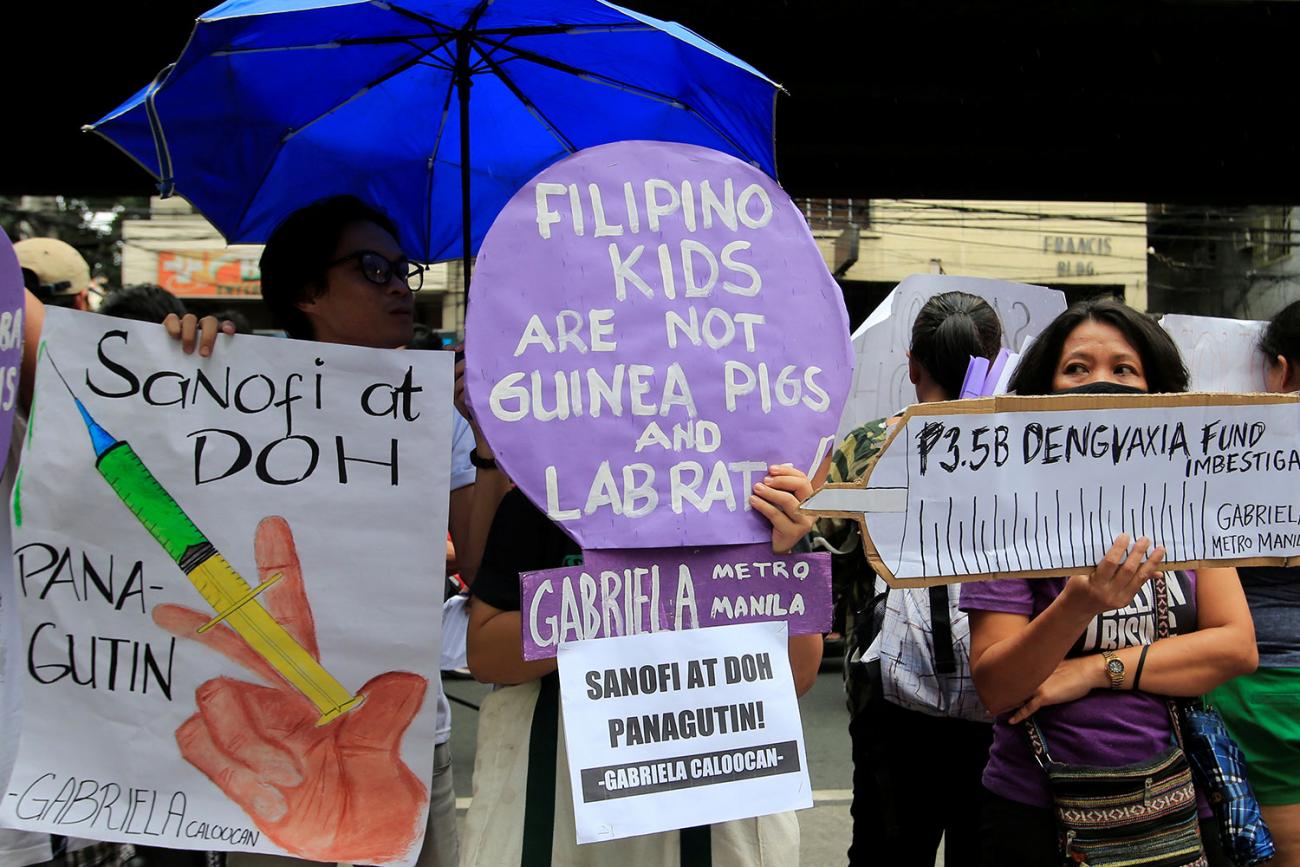 Several protesters are in the picture. Standing with hand-made signs. One holds a sign that reads: "Sanofi and DOH should be held accountable."