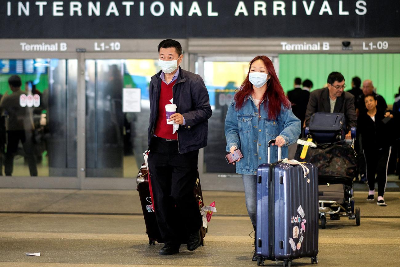 Image shows two travelers with their bags marching out of a gate that says “International Arrivals” in bold letters. They are both wearing dust masks. 