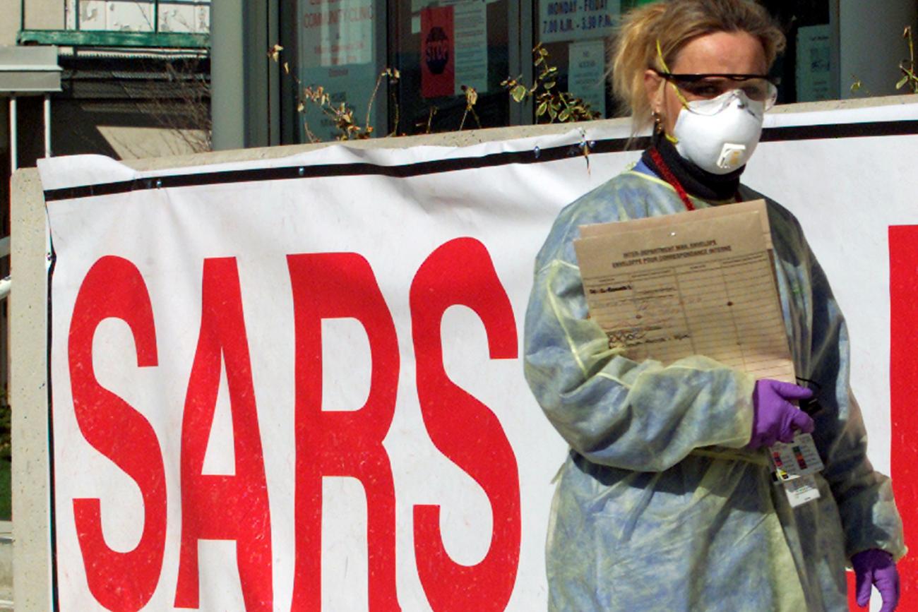 The image shows a woman wearing full protective gear, including eye protection and a surgical mask in front of a hospital adorned with a huge sign that says “SARS” in bright red letters.