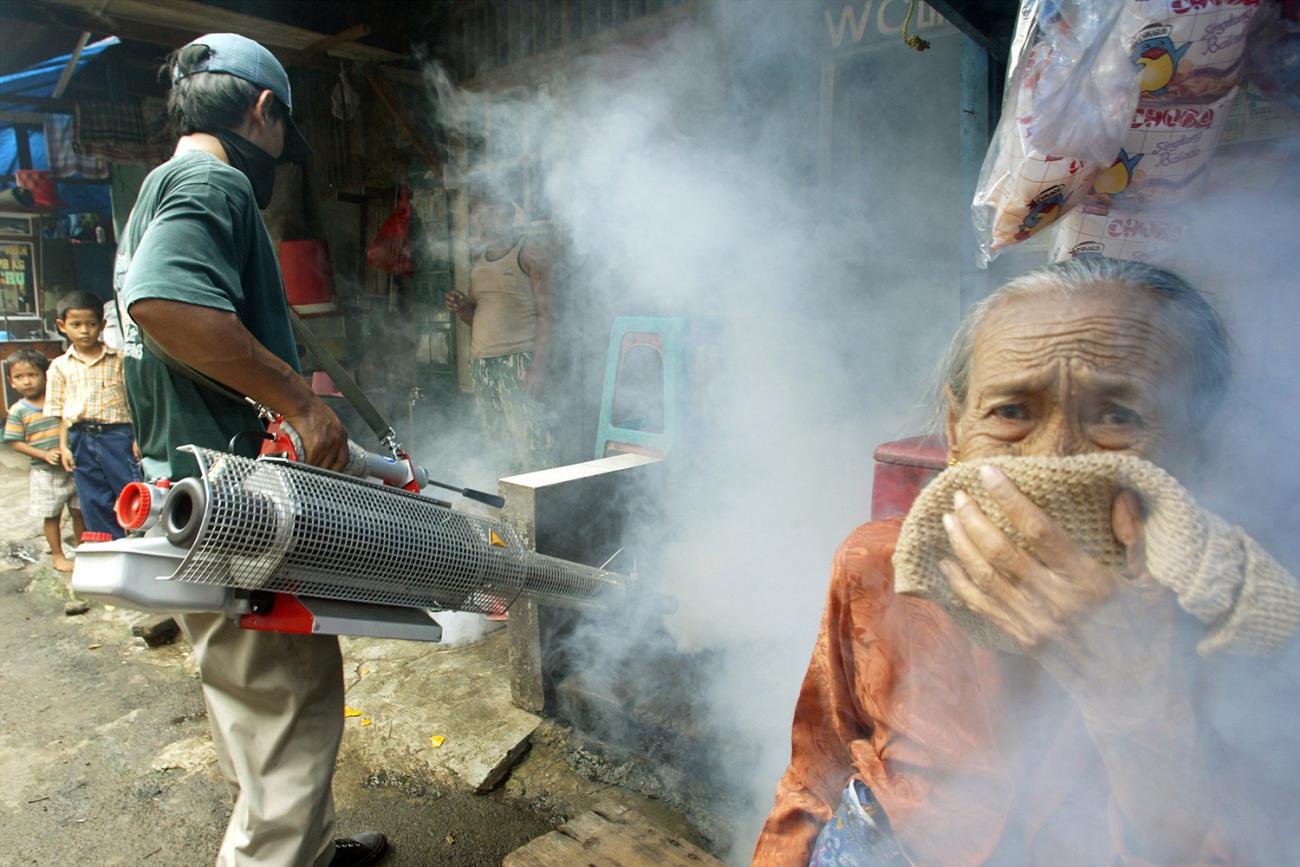 An elderly Indonesian woman covers her nose. The smoke-like bug spray is fogging all around her, and a man can be seen in the background hoisting an industrial spray applicator. This is a striking photo.