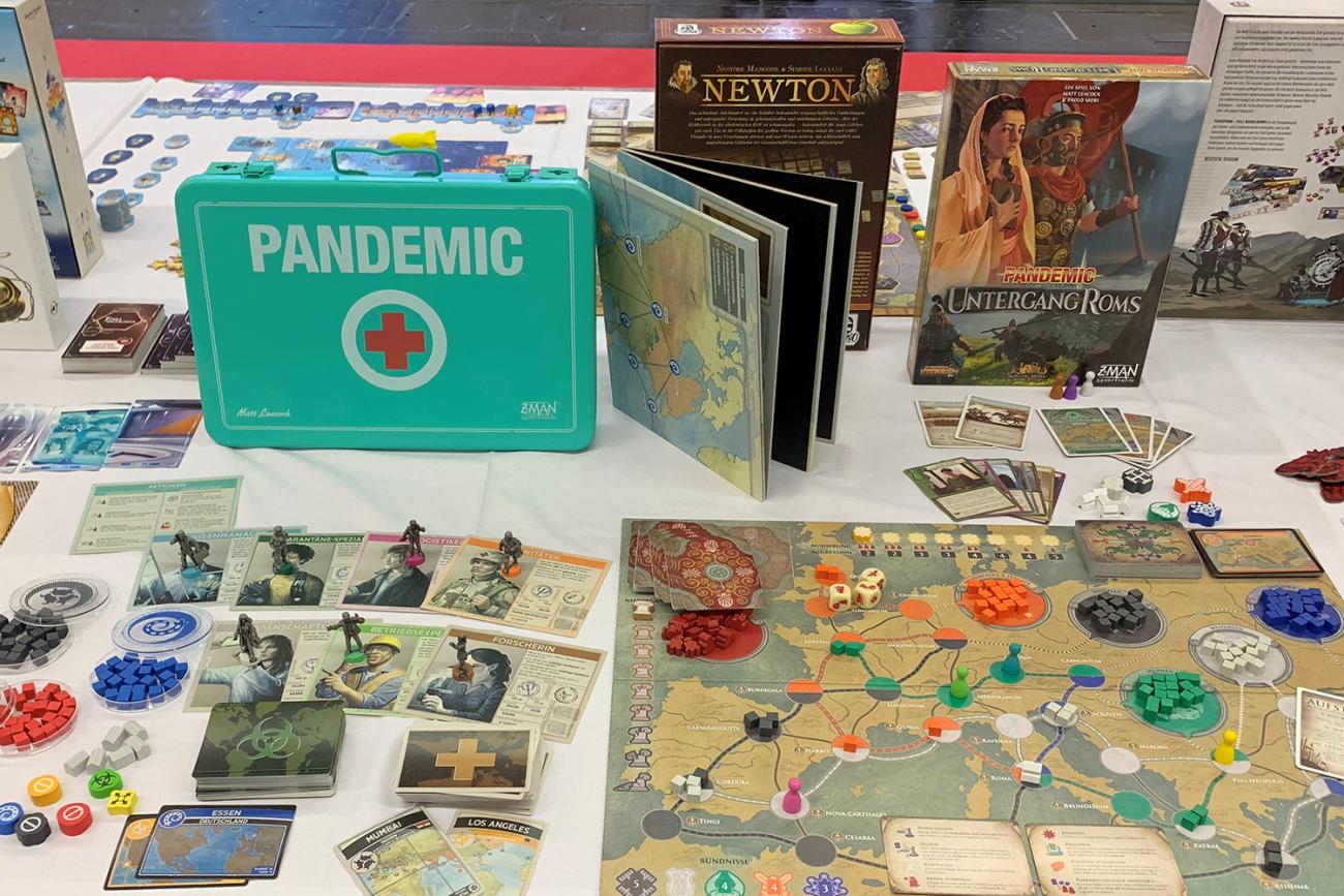 Photo shows a long table on which is sitting a game board showing a map of Western Europe as well as decks of cards, petri dishes of colorful game pieces and several other game boxes on display. 
