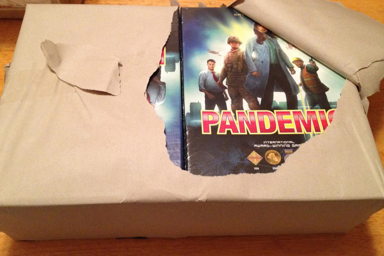 Photo a large armful-sized package neatly wrapped in brown paper. The paper is slightly torn open, revealing eight new boxes of the game Pandemic. 