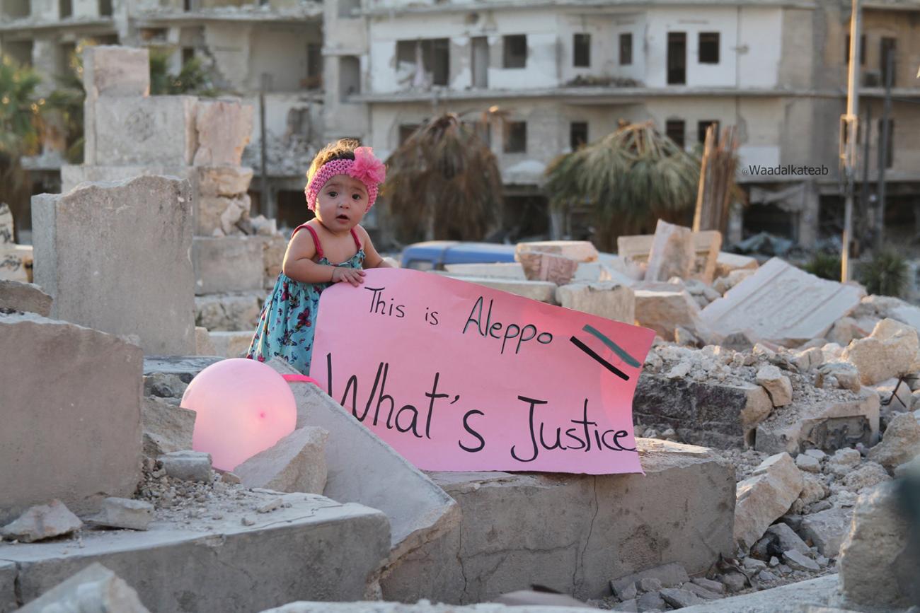 The picture shows the girl, a baby, really, standing on a pile of rubble amidst the destruction of the city holding a sign that reads, “This is Aleppo. What is Justice?” The sign is pink, and there is a pink balloon next to her, and she has a pink head band. 