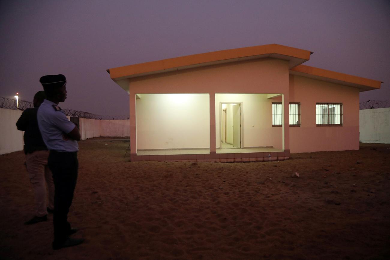Photo shows a very spare, nondescript low-rise building inside a walled compound at dusk. A uniformed guard stands at attention outside. The interior of the building is well lit and there are what appear to be bars on the window.