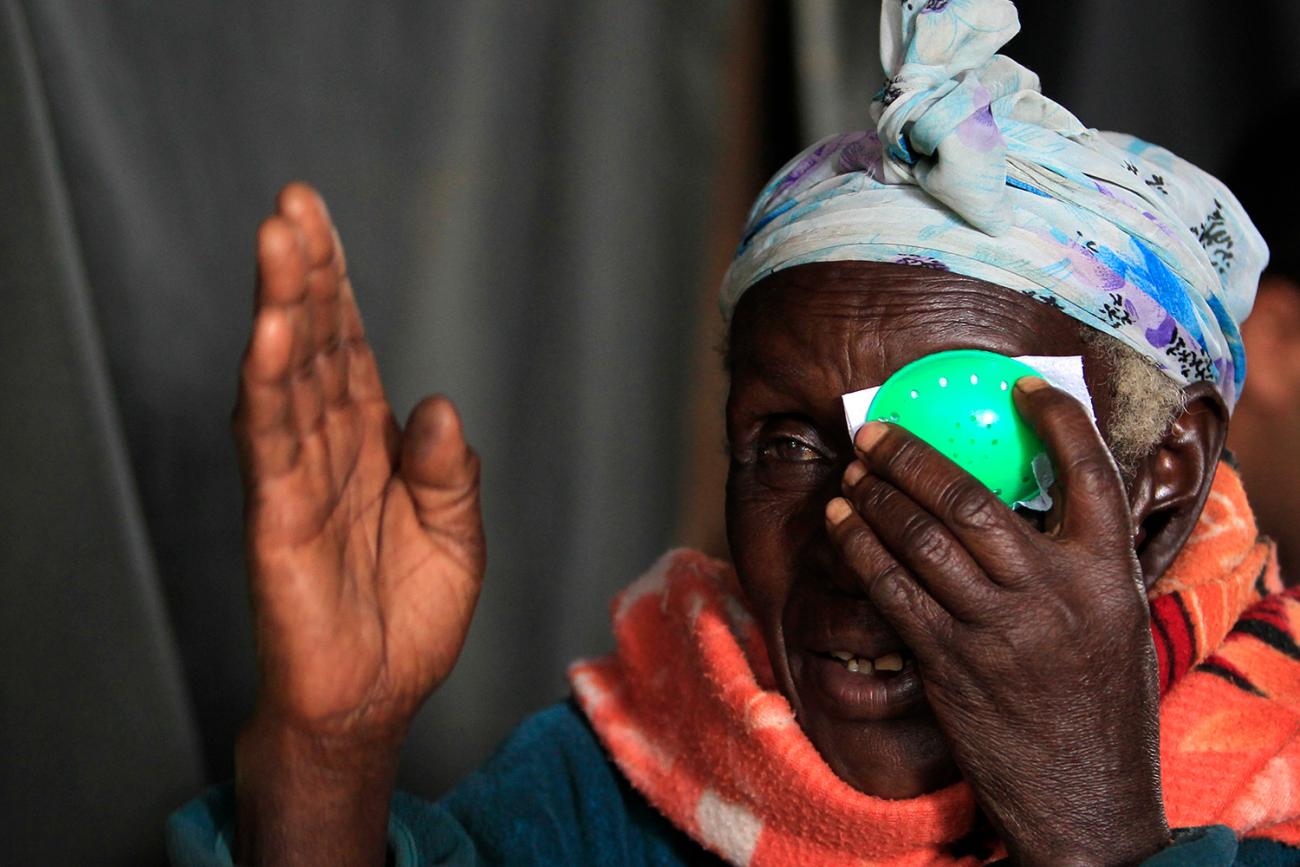 The photo shows a mature woman wearing a bright orange scarf taking an eye test, holding a green patch to her left eye and raising her right hand in front of her face. 
