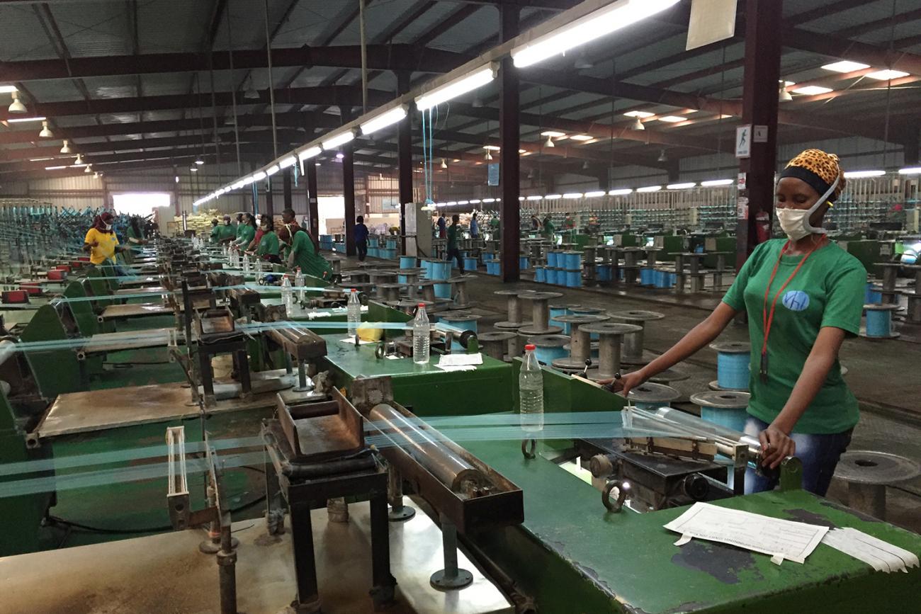The image shows a large factory filled with machines and several workers at the controls of what appear to be industrial looms stretching fabric fibers. 