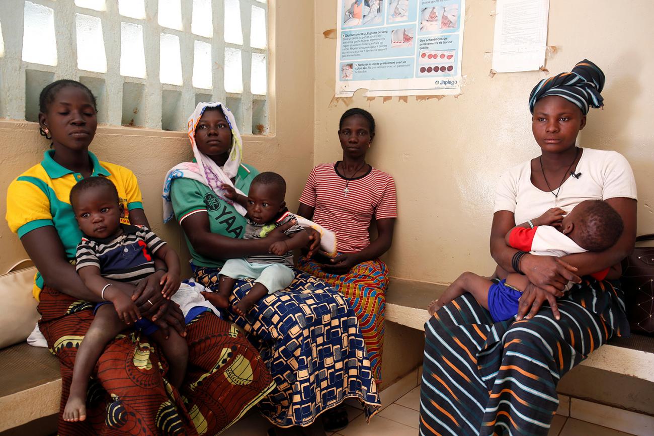 Picture shows four women sitting on a waiting room bench, three of whom have young children on their laps. 