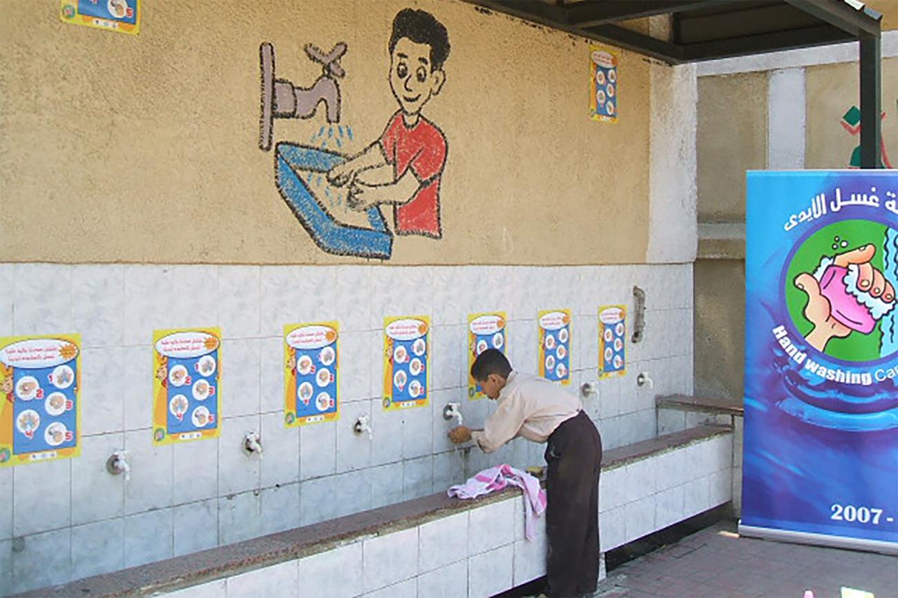 Entitled, “Hygiene Program for Egyptian School Children,” the image shows a boy at a handwadshing station bent over washing his hands. A hygiene propaganda poster can be seen in the backgroung. 