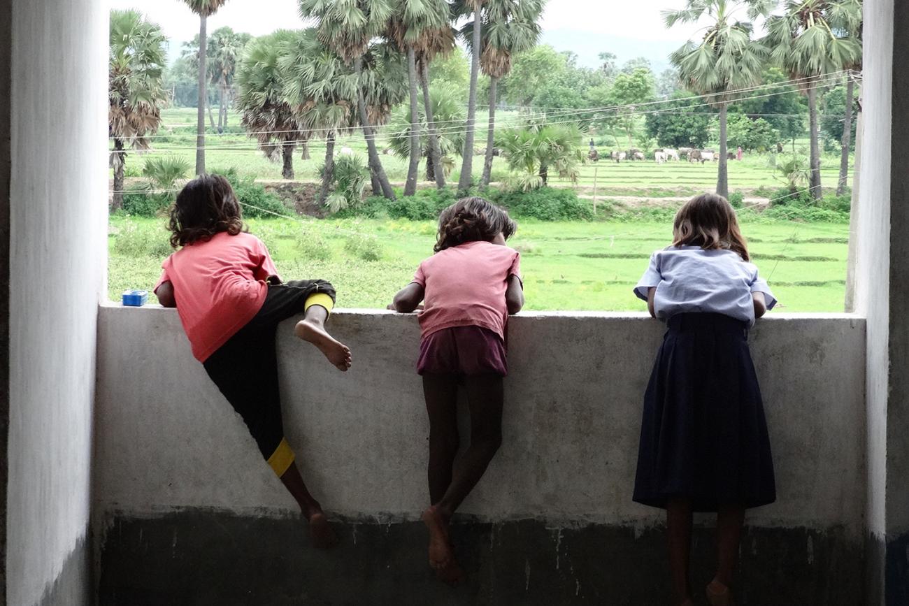 The image shows three children leaning over a wall overlooking green fields and palm trees. 