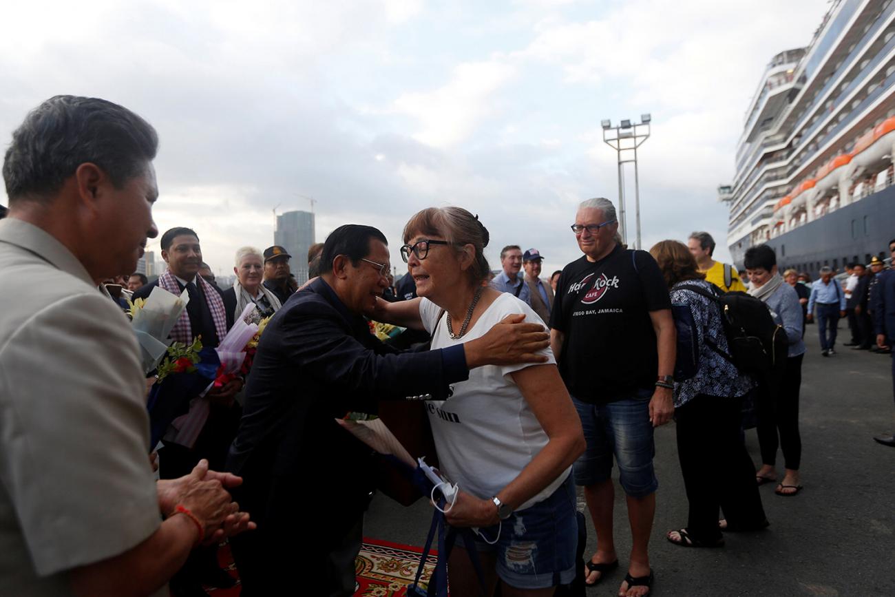 Picture shows the ship docked and disembarked passengers being greeted by the PM. He is going in for a hug with one lady. 