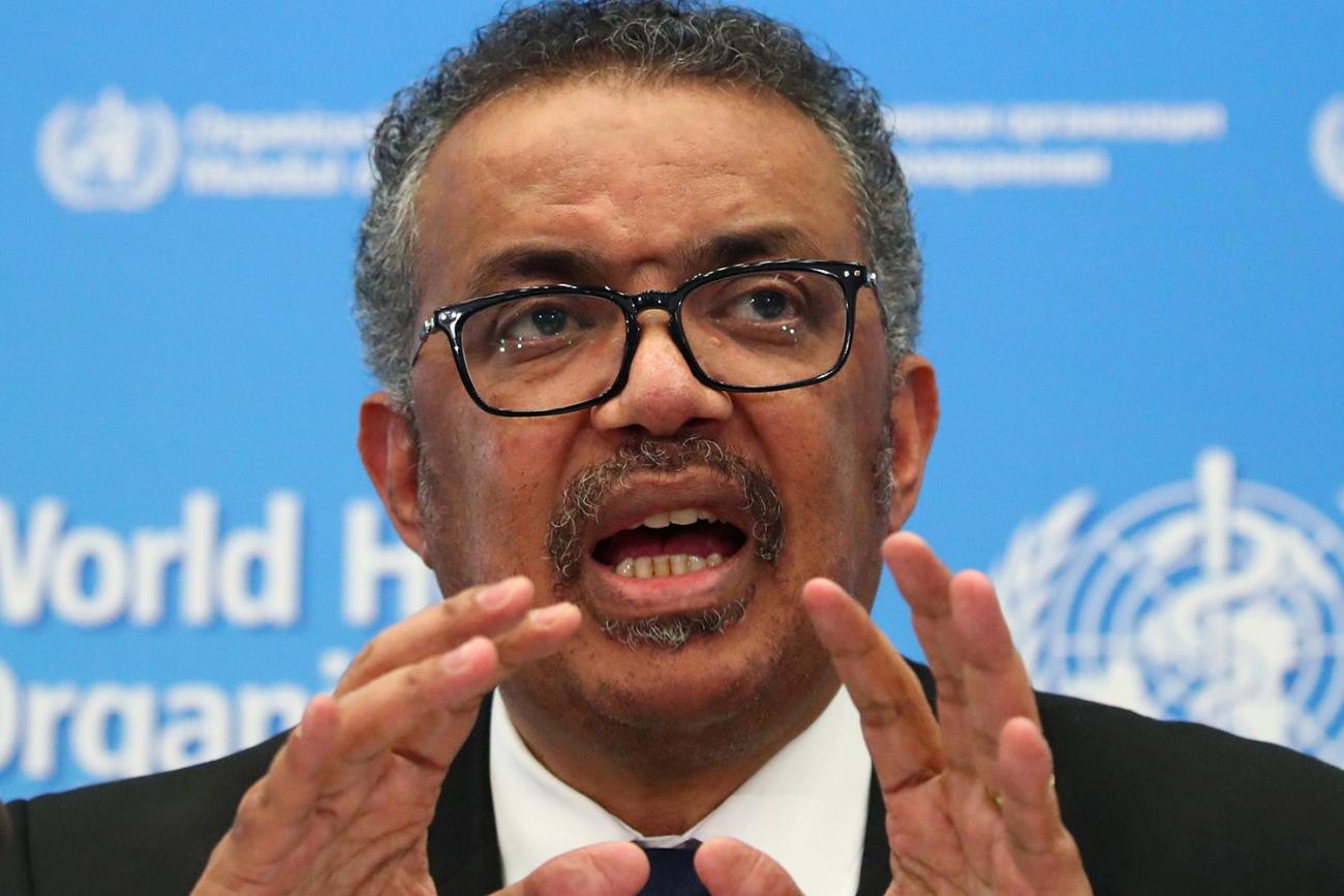 Photo shows the Director-General of the WHO speaking at a table at a press conference and gesturing with his hands. 