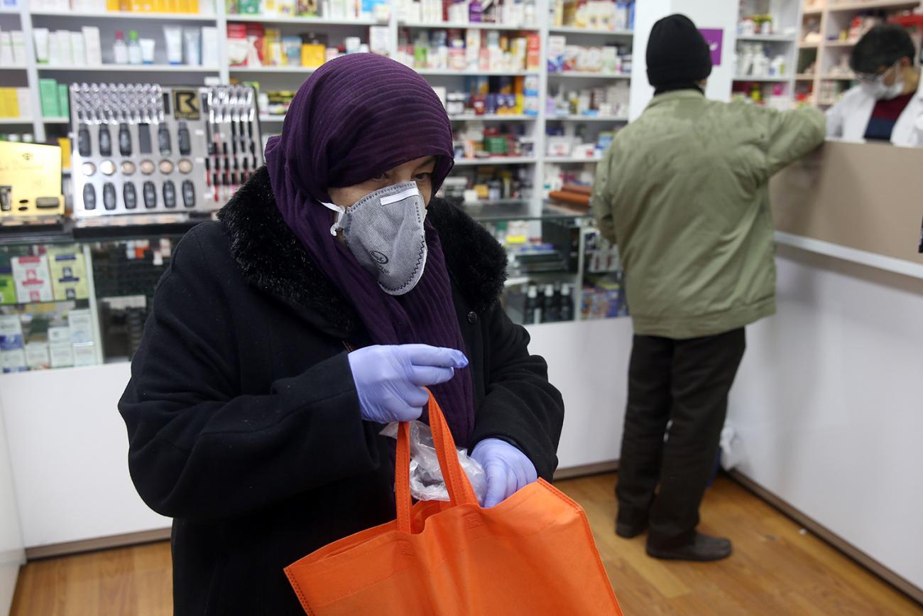The photo shows a woman appearing to be leaving the drug store holding a bright orange bag with purple gloves on. In the background the pharmacist can be seen halping another customer. 