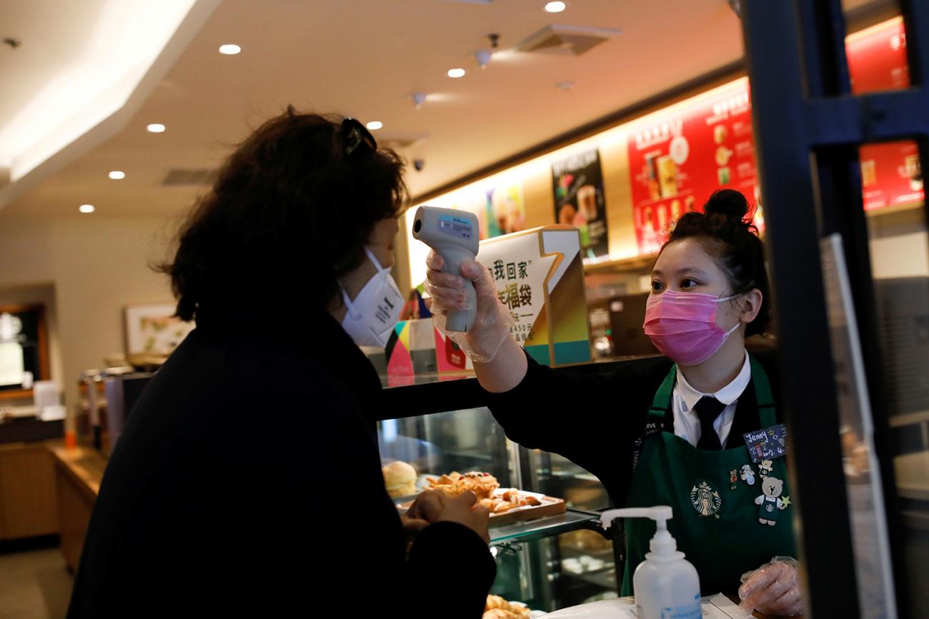 The photo shows a counter worker taking the temperature of a customer with an infrared gun-type thermometer. Both are wearing masks. 
