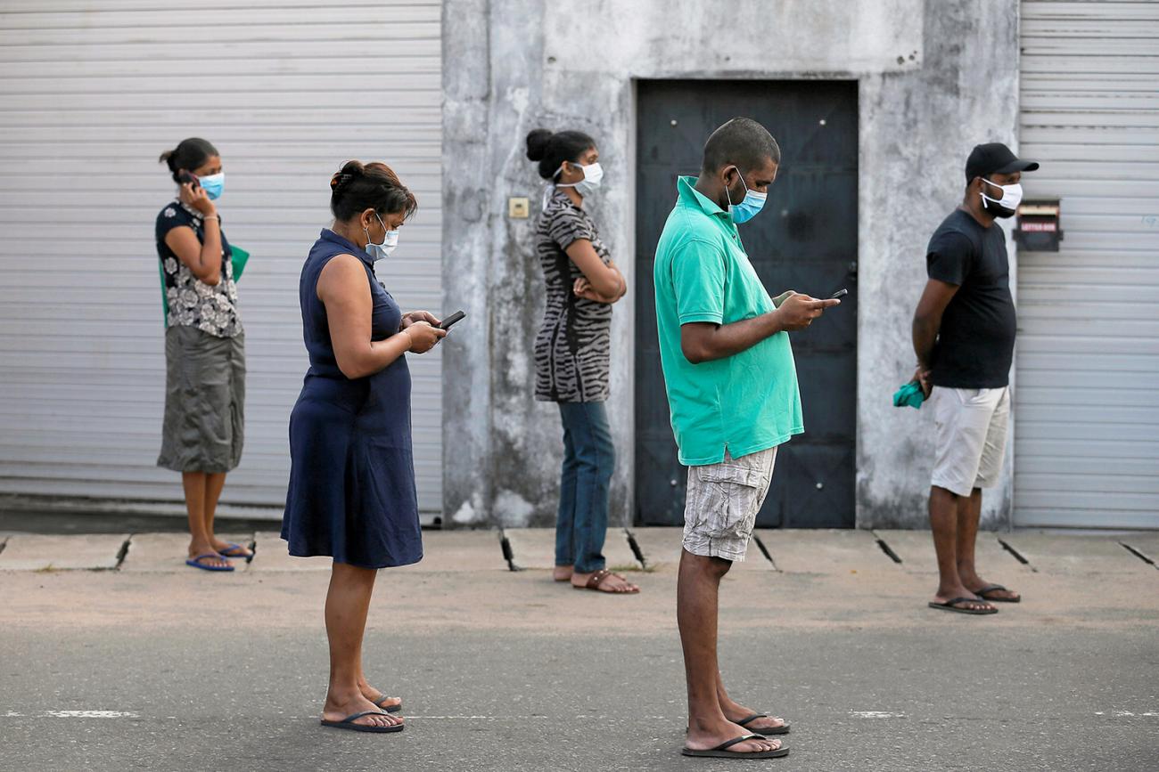 The image shows a line of people in warm weather clothing waiting in line. Most of them are wearing masks. 