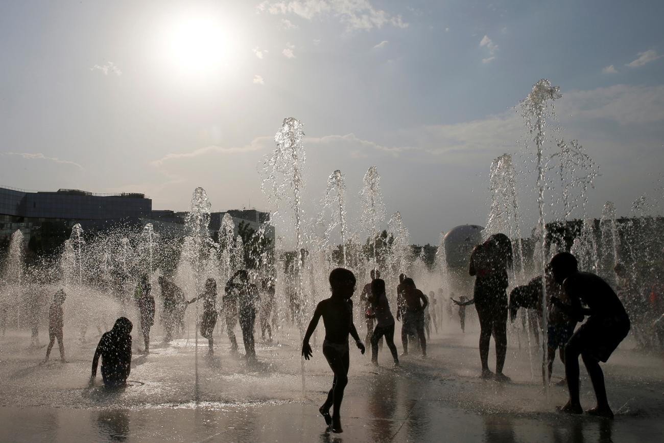 The picture shows a number of children silhouetted as they cool off in water fountains at an urban park. 