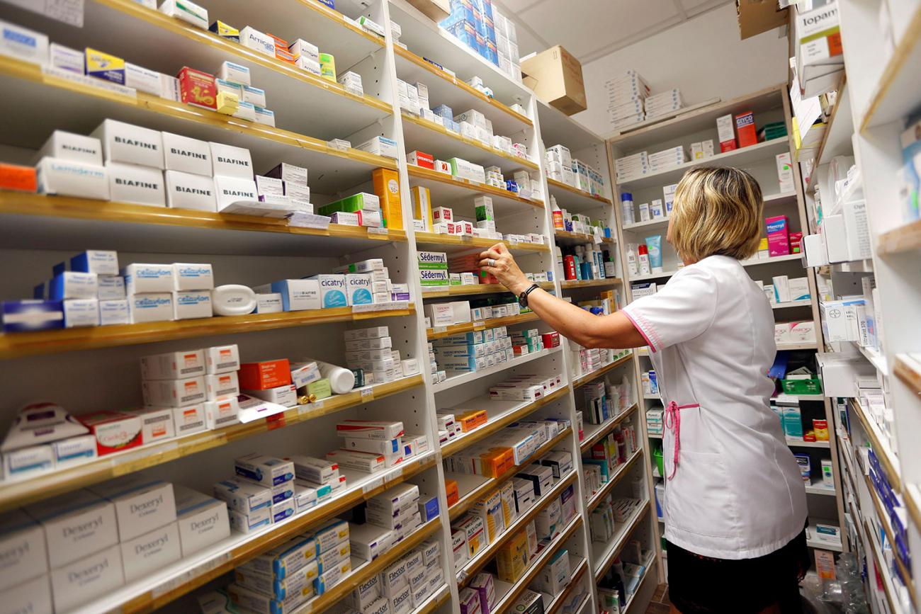 The photo shows a pharmacist with a white coat standing in front of a shelf filled with numerous medicines. 