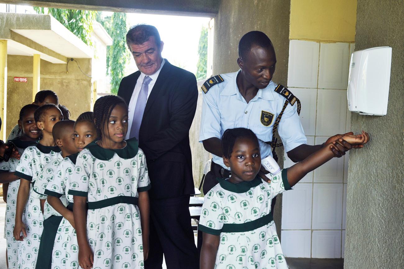 Picture shows a line of girls in school uniforms queueing and being helped at a hand sanitizer station. 