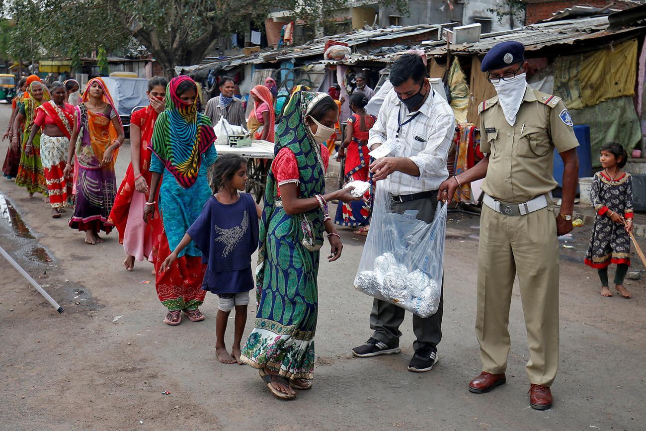 A long line of people, mostly women, stretches off into the distance as two uniformed soldiers wearing masks reach into a plastic bag and pull out foil-wrapped pouches for those in line.