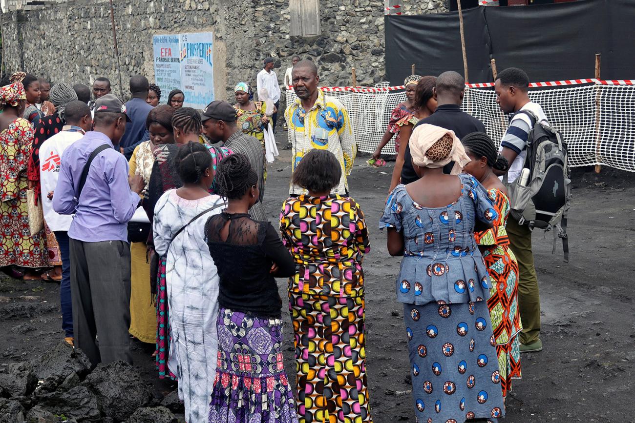 The photo shows many people milling about and forming a crowd around one health worker during daytime. 