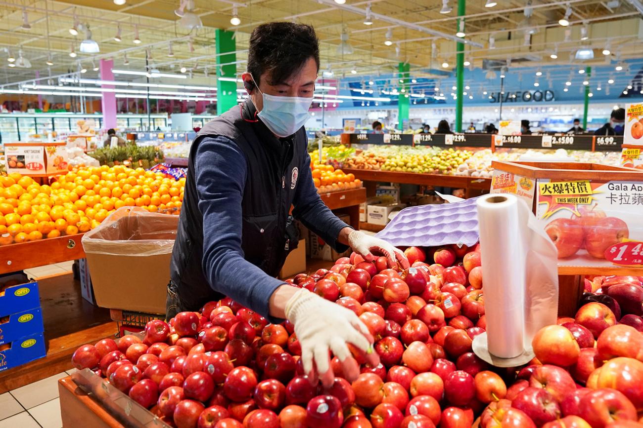 The photo shows a grocery employee stacking apples in a colorful display in a produce section. 