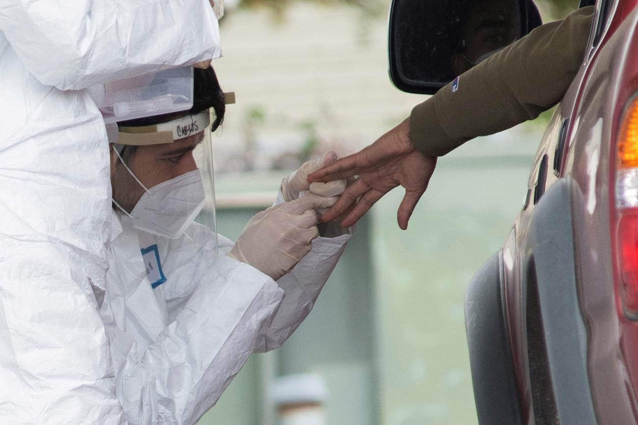 The photo shows a hand reaching out of a car while a health worker knees beside the vehicle administering the test. 