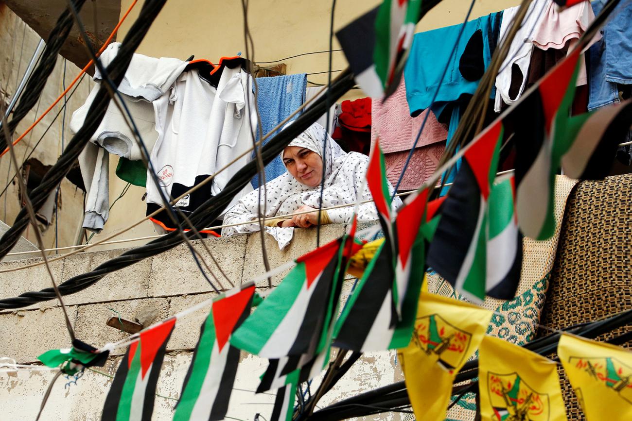 Picture shows the woman looking down at the camera from a balcony above. Clothes strewn on a line behind her and a string of Palestinian flags hung between her and the camera visually mix to make the scene very busy with billowing cloth. 
