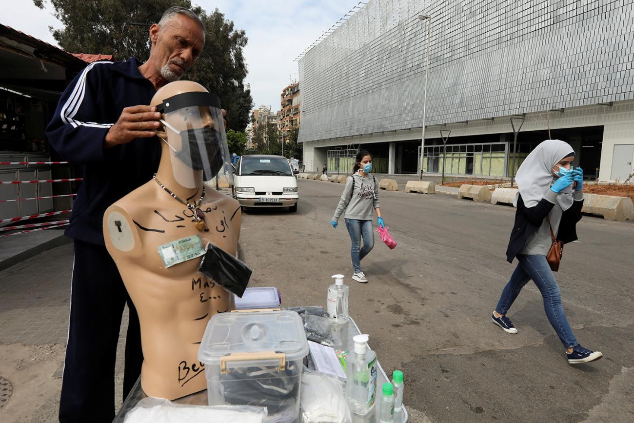  The photo shows the man fitting a clear plastic mask on a manikin next to a makeshift huckster table on a busy street with people walking by. 