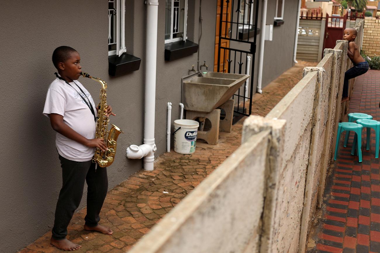 The photo shows a teenage boy playing a huge sax while another, younger boy scambles up over a fence. 