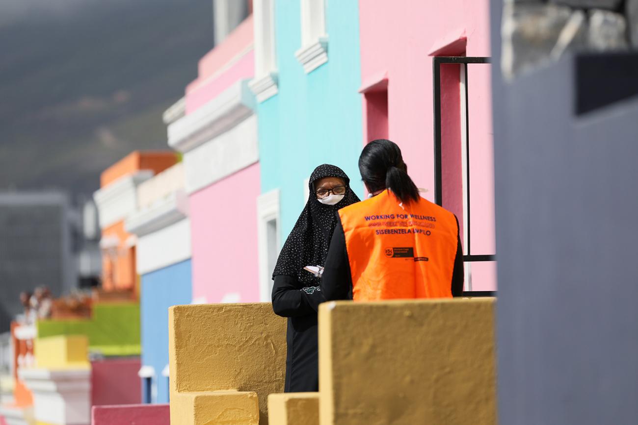 The photo shows the health care worker from behind speaking to a women wearing a head scarf and mask and facing the camera. They are standing on the front steps of a series of houses that are brightly colored green and orange and pink and purple pastels. 