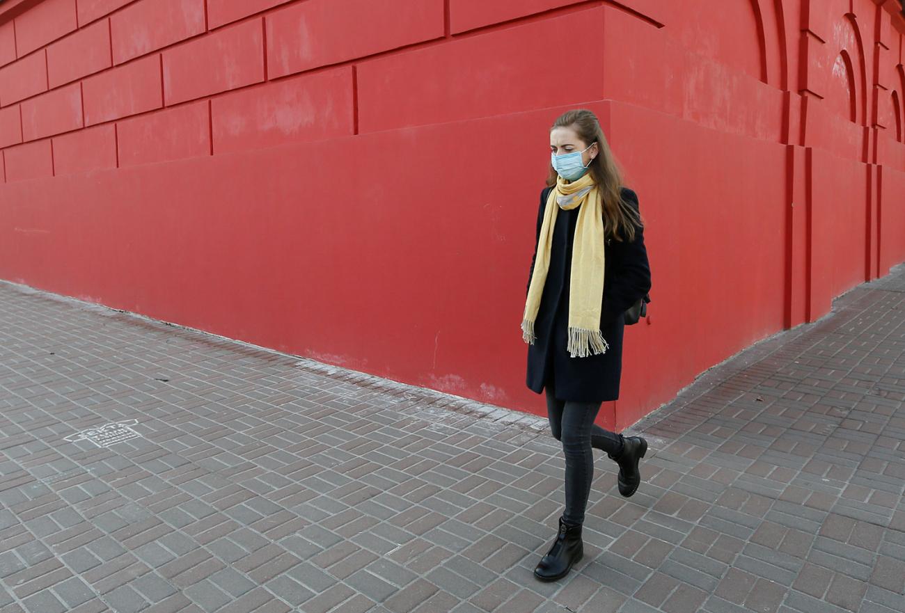  The photo shows a young woman wearing a face mask walking down a sidewalk with a massive building behind her painted brick red. 