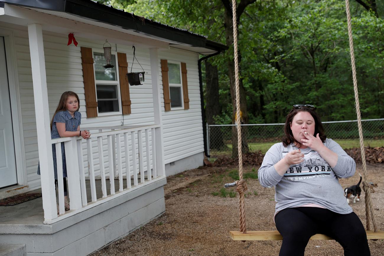 Picture shows the mother sitting on a swing smoking while her daughter stands behind her on the porch. 