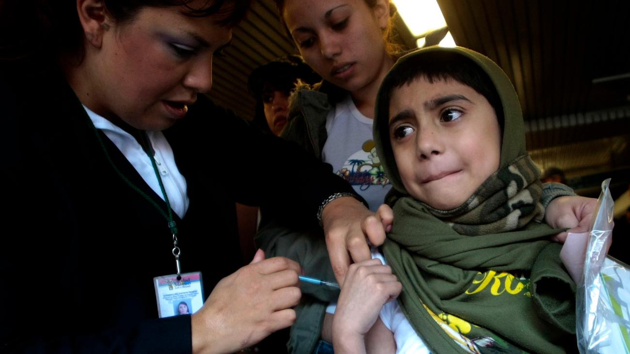 The photo shows a girl getting a shot in a bustling metro station with her mother looking on. 