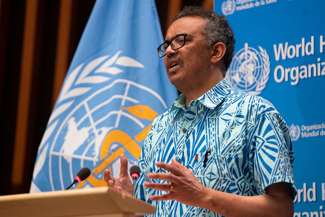 The WHO official is at a podium speaking, and surprisingly wearing a Hawaiian shirt.
