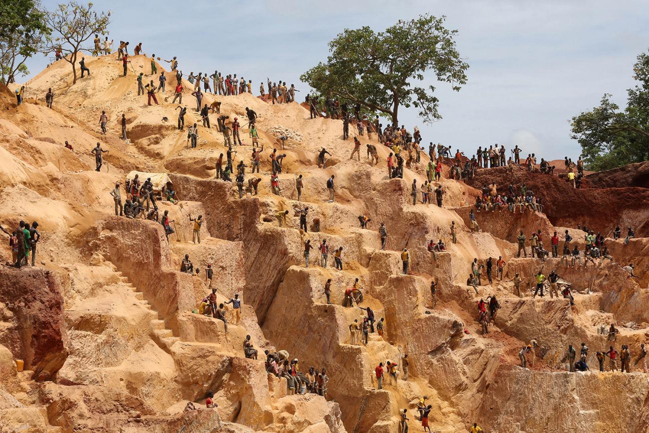Picture shows the steppes of a small mountain-sized mine cut into the red earth with tiny figures of people everywhere on the different levels. 