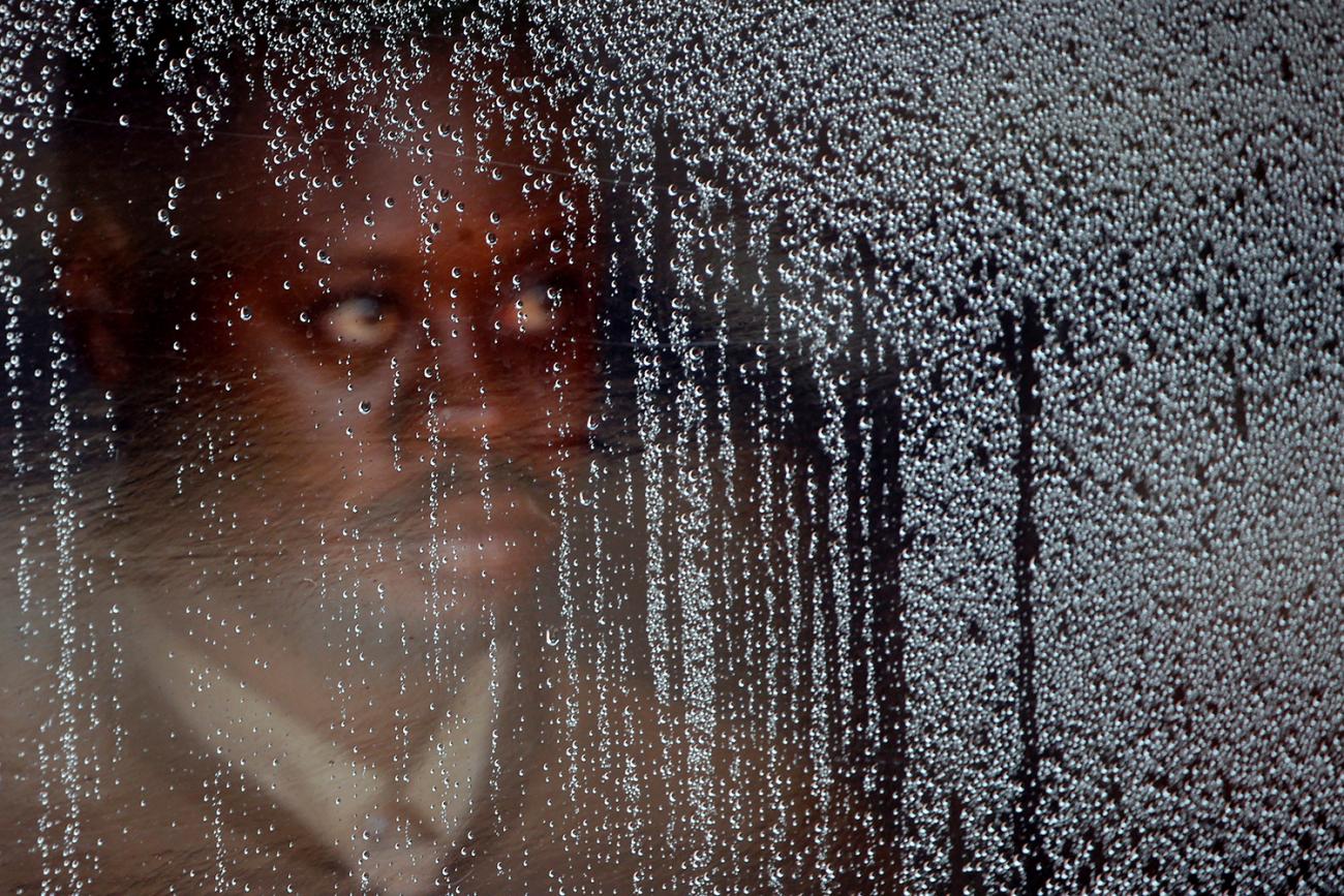 Picture shows the man looking out behind the clear plastic screen, which is covered with water droplets, presumably from rain. 