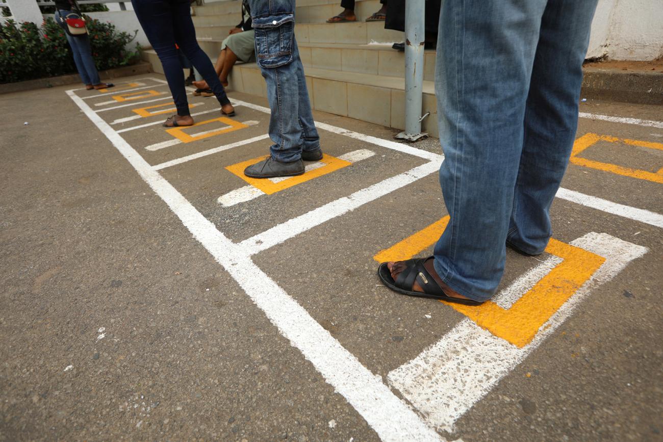 Photo shows the legs of several people standing in social distancing squares painted on a blacktop as they wait in line. 