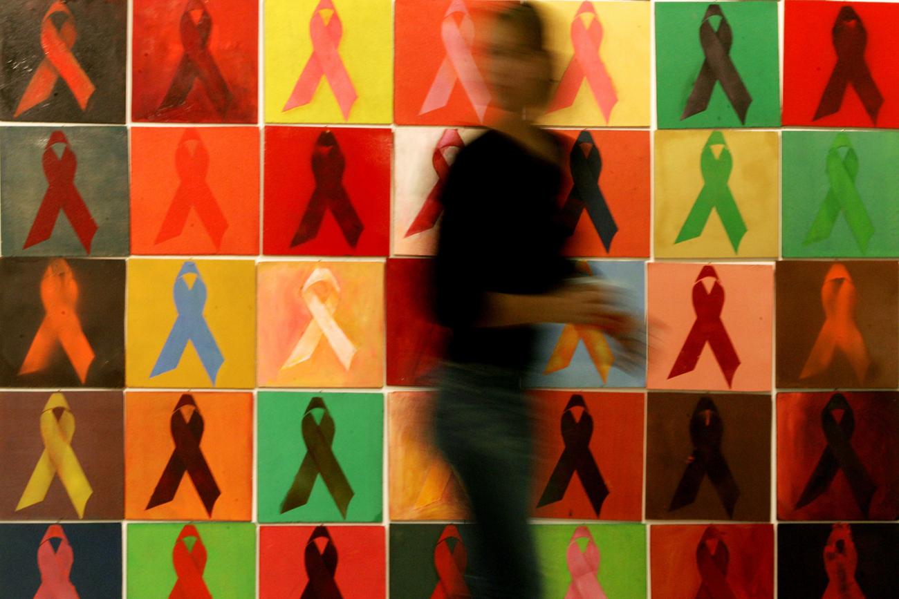The photo shows a blurred figure walking past a mural with lots of multi-colored AIDS ribbons, reminiscent of an Andy Warhol. 