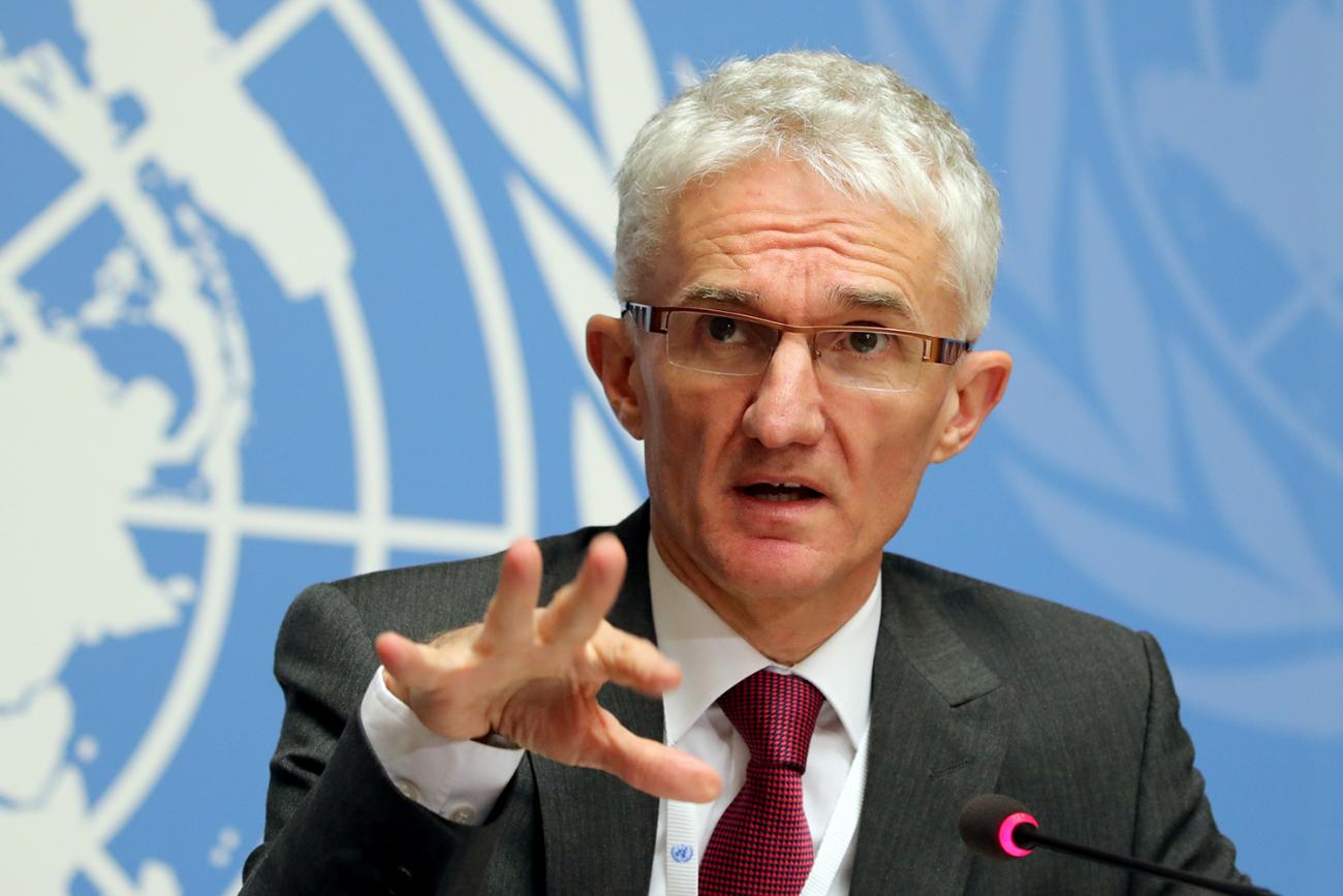The official is seated at a table speaking into a microphone and gesturing with his hand. A blue and white UN logo is behind him. 