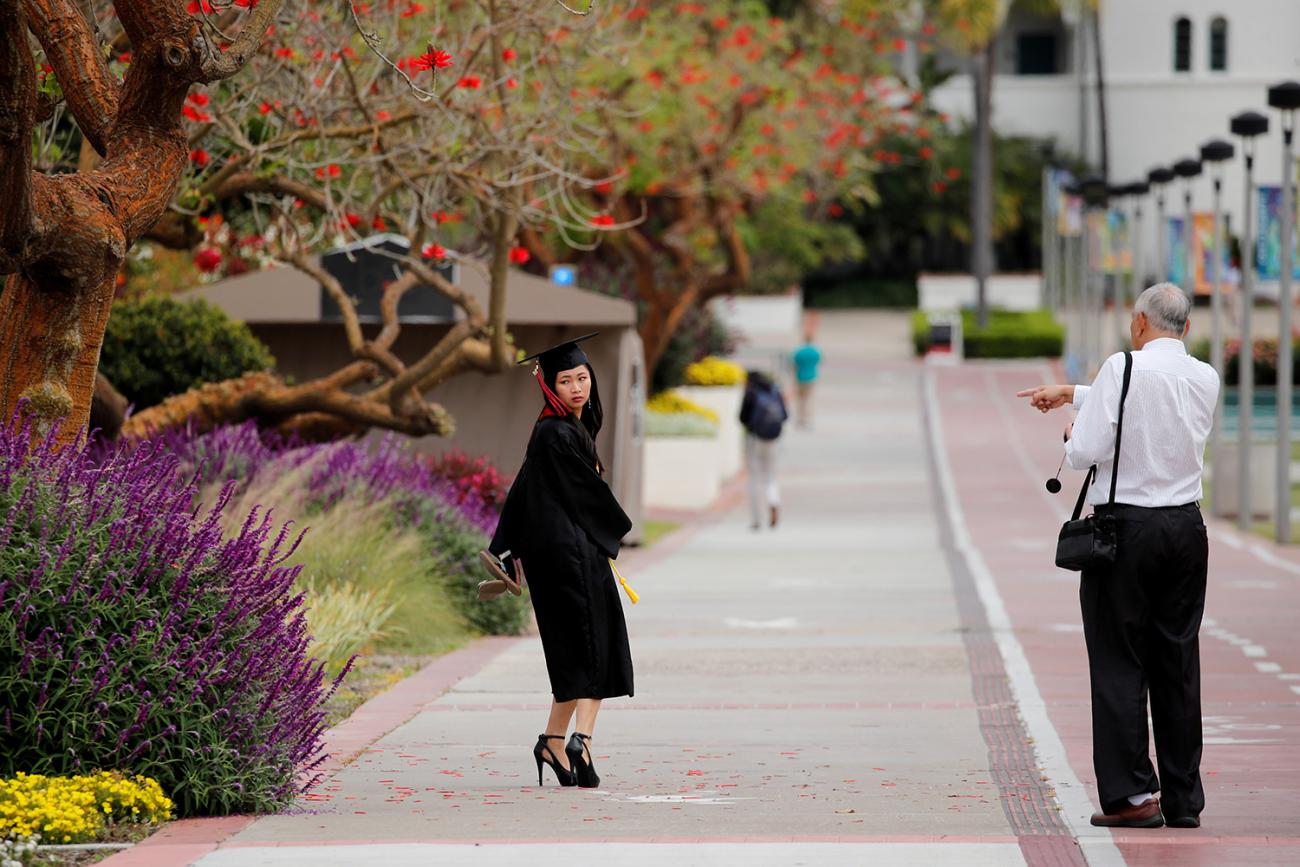 The photo shows a well-dreSsed graduate in a cap and gown posing by a lush tropical landscape. 