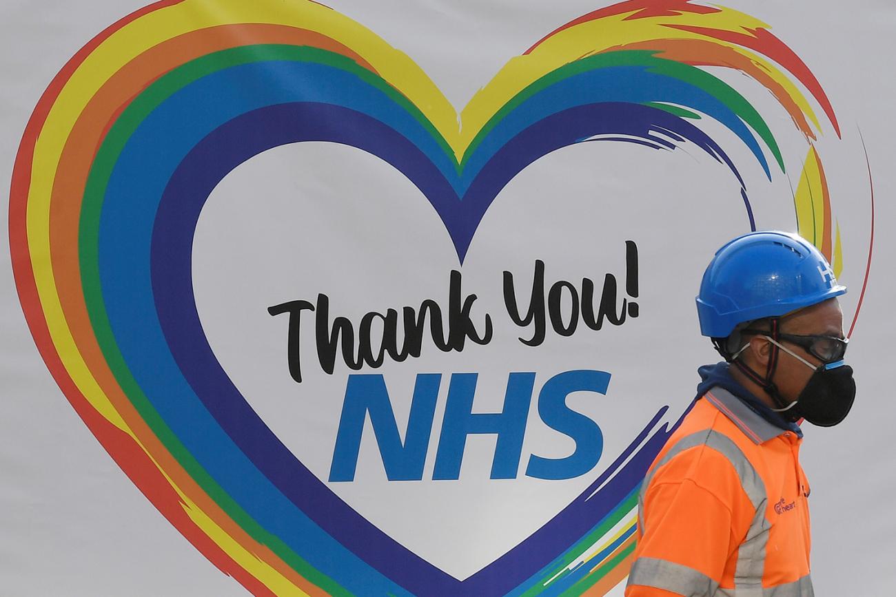 The photo shows a construction worker with a hardhat and face mask walking past a sign that reads "Thank You! NHS" and is surrounded by a rainbow heart. 