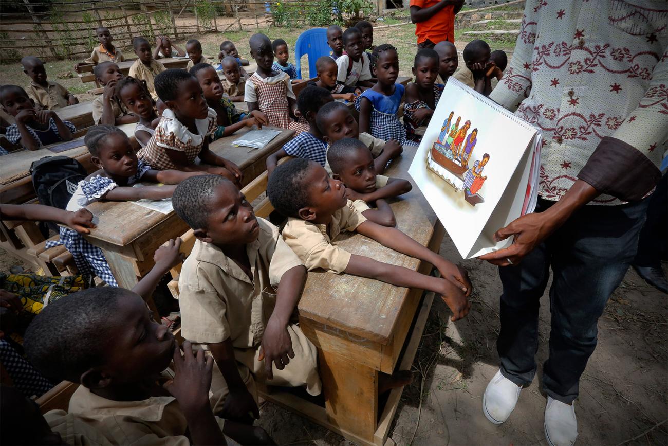 The photo shows a man in front of a class full of kids holding an instructive placard. 