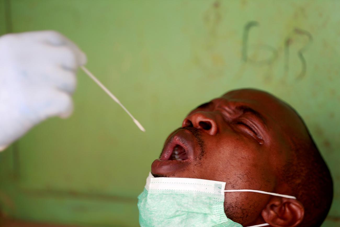 The photo shows a man with his head bac, a medical worker holding a long swab in his gloved hand. The man looks stressed, though perhaps he is just reacting to the swabbing. The background is lime green. This is a dramatic photo. 