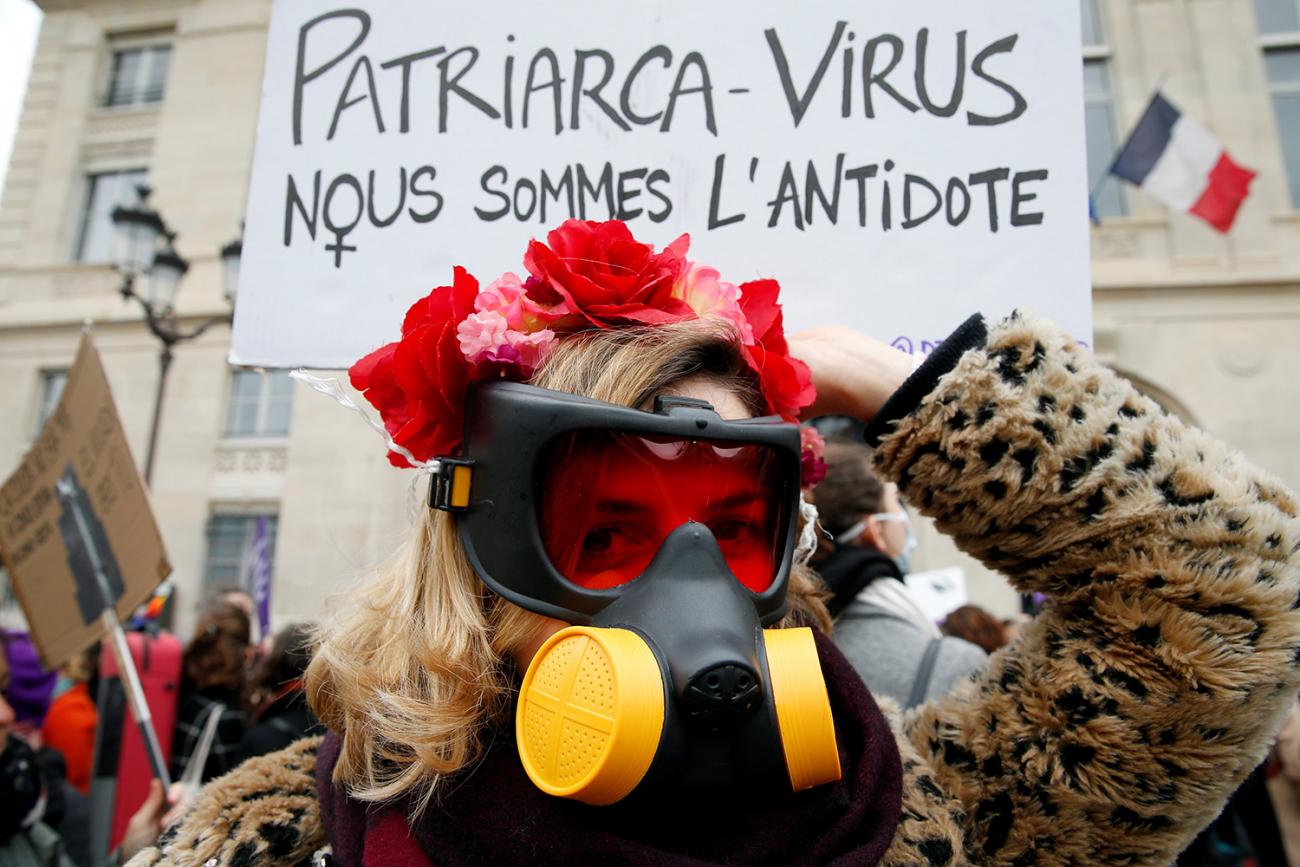 The woman is wearing a gas mask and stands in front of a placard reading, in French, "Patriarchy virus. We are the antidote."