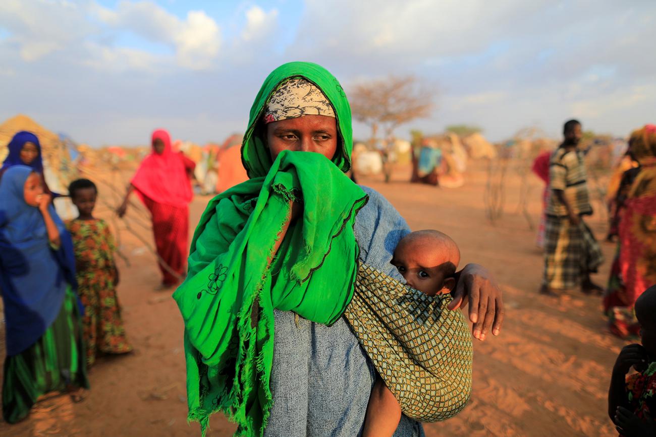 The photo shows a woman with a bright green scarf holding a small child bundled to her torso. She is covering her face and has a look of extreme sadness on her face. The child looks frightened. 